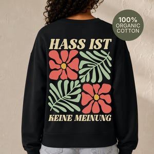 Könnte beinhalten: Ein schwarzes Langarm-Sweatshirt aus 100 % Bio-Baumwolle. Auf der Rückseite befindet sich ein Aufdruck mit dem Text HASS IST KEINE MEINUNG, ergänzt durch ein Gittermuster aus zwei korallenfarbenen Blumen und zwei hellgrünen Farnblatt-Illustrationen.