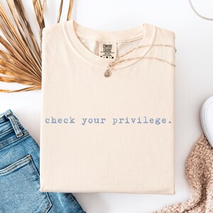 Può includere: T-shirt color crema con la scritta "check your privilege" in azzurro. La maglietta è piegata e presentata con jeans blu, scarpe da ginnastica bianche e una delicata collana. Una foglia di palma secca aggiunge un tocco naturale.