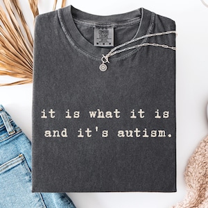Könnte beinhalten: Ein anthrazitfarbenes T-Shirt mit Rundhalsausschnitt und einem Comfort Colors Markenetikett am Innenkragen. Das Shirt zeigt den weißen Schriftzug "it is what it is and it's autism." in einer Schreibmaschinen-Schriftart mittig auf der Brust.