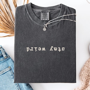 Peut inclure: T-shirt gris foncé avec l'inscription blanche « stay weird ». Un collier argenté avec un pendentif soleil est posé sur le t-shirt plié. Un jean bleu et une chaussure blanche sont visibles à côté.