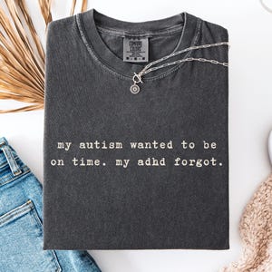 Könnte beinhalten: Ein anthrazitfarbenes Kurzarm-T-Shirt mit dem in Weiß aufgedruckten Text "my autism wanted to be on time. my adhd forgot.". Das T-Shirt hat einen Rundhalsausschnitt und ein Comfort Colors Markenetikett am Innenkragen, flach ausgelegt.