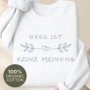Può includere: Una felpa bianca a maniche lunghe con il testo blu HASS IST KEINE MEINUNG e un grafico centrale di due piccoli cuori affiancati da rami frondosi. Un badge circolare sul capo indica 100% ORGANIC COTTON, colore bianco.