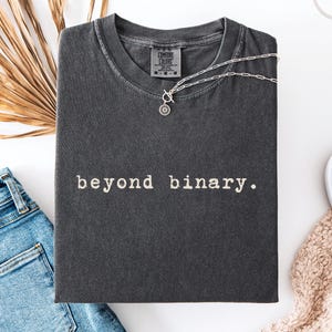 Könnte beinhalten: Ein pfefferfarbenes T-Shirt mit Rundhalsausschnitt und dem Text "beyond binary.", der in einer Schreibmaschinen-Schriftart auf der Brust gedruckt ist. Das Shirt verfügt über ein Comfort Colors Markenetikett am inneren Halsausschnitt.