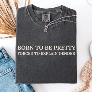 Könnte beinhalten: Ein anthrazitfarbenes T-Shirt mit Rundhalsausschnitt und dem in hellbeiger Schrift gedruckten Text BORN TO BE PRETTY FORCED TO EXPLAIN GENDER. Das Shirt verfügt über ein Comfort Colors Markenetikett am Innenkragen und ein verwaschenes Vintage-Finish.