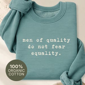 Könnte beinhalten: Ein langärmeliges, blaugrünes Sweatshirt mit Rundhalsausschnitt und dem weißen Text "men of quality do not fear equality." Ein kreisförmiges Abzeichen unten links zeigt "100% ORGANIC COTTON" und oben rechts steht "COLOR: GREEN BAY."