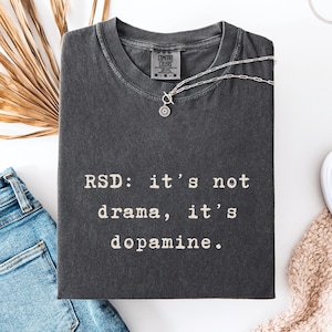 Könnte beinhalten: Ein anthrazitfarbenes Kurzarm-T-Shirt mit Rundhalsausschnitt und einem Comfort Colors Markenetikett. Das T-Shirt zeigt den weißen aufgedruckten Text: RSD: it's not drama, it's dopamine. Eine silberfarbene Kette mit einem runden Anhänger liegt am Kragen.