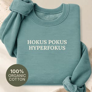 Op de afbeelding: Een groen bay gekleurd sweatshirt met lange mouwen en de witte tekst HOKUS POKUS HYPERFOKUS op de borst gedrukt. Een cirkelvormige badge in de linkerbenedenhoek toont de tekst 100% ORGANIC COTTON. Het kledingstuk heeft een ronde halslijn.