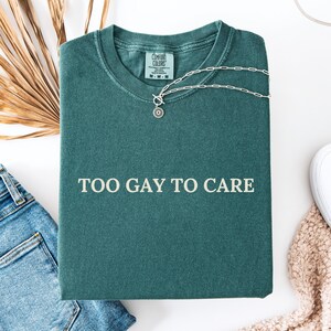 Può includere: Una maglietta blu abete con la scritta bianca "TOO GAY TO CARE". La maglietta è piegata e ha una collana d'argento con un ciondolo circolare. Un paio di jeans blu e una scarpa bianca sono visibili sullo sfondo.