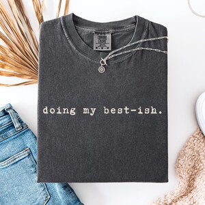 Könnte beinhalten: Gefaltetes, pfefferfarbenes T-Shirt mit dem weißen Schriftzug „doing my best-ish“. Eine silberne Halskette mit Sonnenanhänger liegt auf dem Shirt. Blaue Jeans und ein weißer Schuh sind ebenfalls zu sehen.