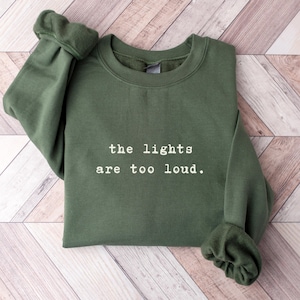 May include: An olive green sweatshirt with the phrase "the lights are too loud" printed in white. The sweatshirt is made of a soft material and has a crew neck and long sleeves.