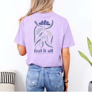 Puede incluir: Camiseta lila con un dise&ntilde;o floral en la espalda. El dise&ntilde;o incluye flores azules y grises con las palabras "feel it all" y "every emotion has its place" impresas debajo.