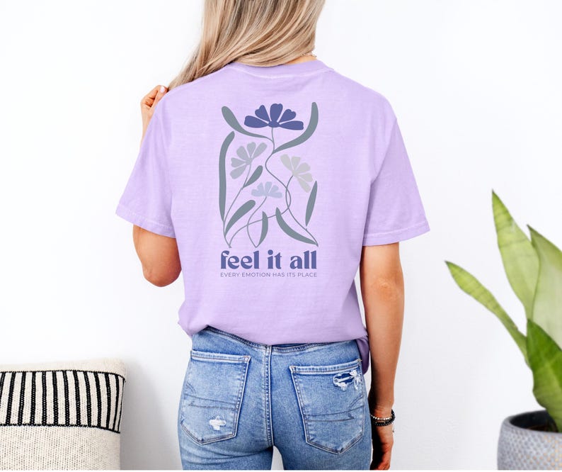 Puede incluir: Camiseta lila con un dise&ntilde;o floral en la espalda. El dise&ntilde;o incluye flores azules y grises con las palabras "feel it all" y "every emotion has its place" impresas debajo.