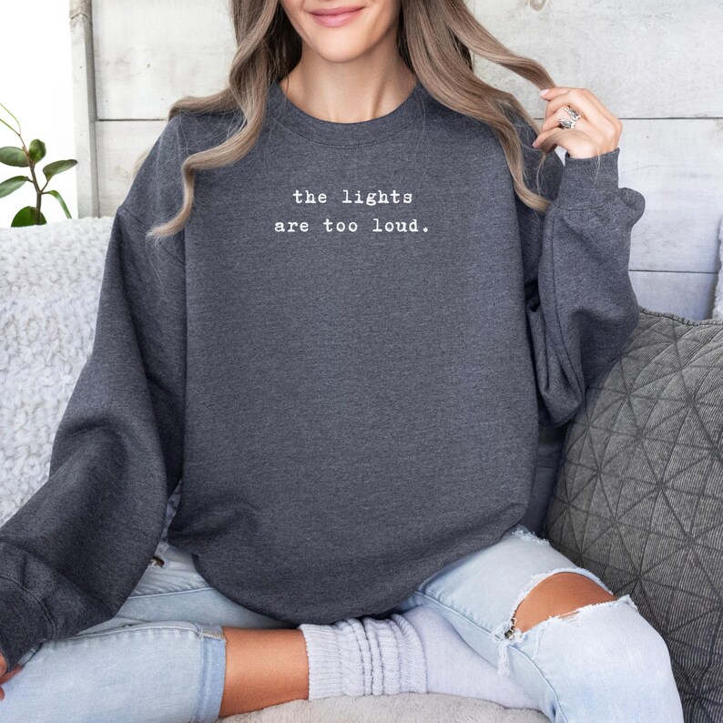 May include: Dark gray sweatshirt with the text "the lights are too loud" in white. The sweatshirt has long sleeves and a crew neck. The model is wearing light blue jeans with ripped knees.