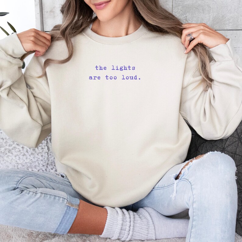 May include: Cream-colored sweatshirt with the text "the lights are too loud" in purple. The sweatshirt is a neutral color and has a crew neck. The text is centered on the chest.