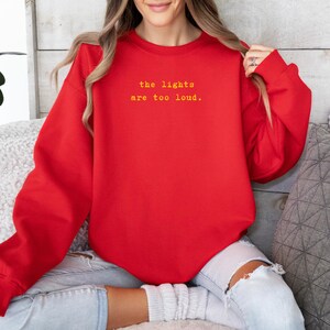May include: A red sweatshirt with the text "the lights are too loud" in yellow. The sweatshirt is a solid color and has a crew neck. The image shows a person wearing the sweatshirt.