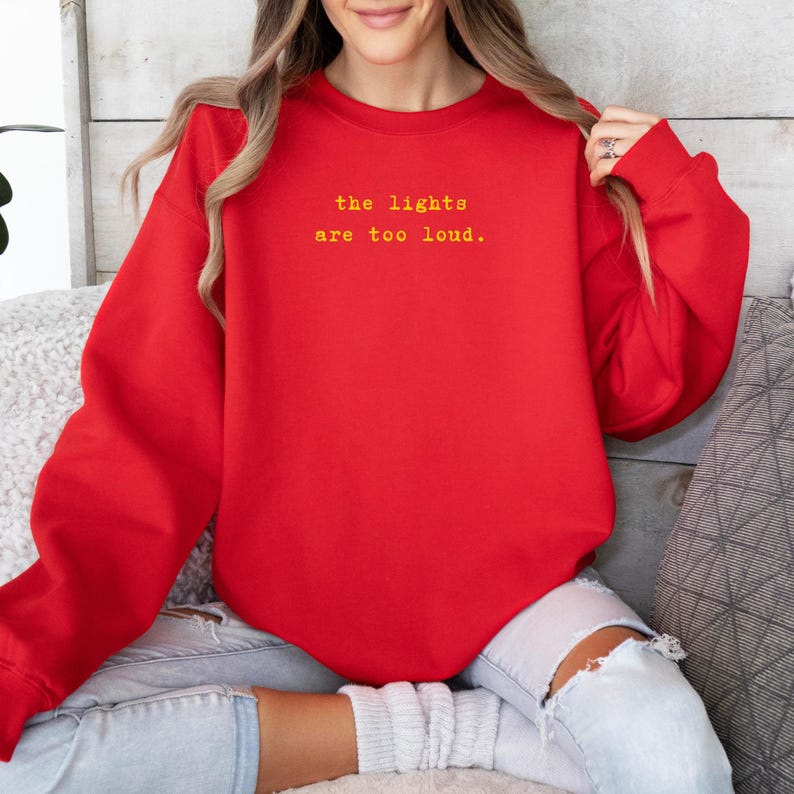 May include: A red sweatshirt with the text "the lights are too loud" in yellow. The sweatshirt is a solid color and has a crew neck. The image shows a person wearing the sweatshirt.