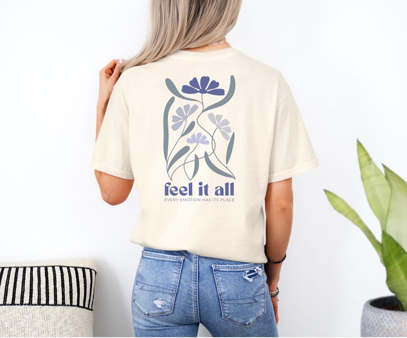 Puede incluir: Camiseta color crema con un dise&ntilde;o floral en la espalda. El dise&ntilde;o presenta flores azules y grises con el texto "feel it all" y "every emotion has its place" en azul. La camiseta est&aacute; hecha de un material suave.