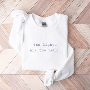May include: A white sweatshirt with the text "the lights are too loud" in light blue. The sweatshirt is folded on a wooden surface with a light brown and white pattern. The Gildan brand tag is visible.