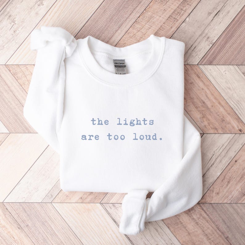 May include: A white sweatshirt with the text "the lights are too loud" in light blue. The sweatshirt is folded on a wooden surface with a light brown and white pattern. The Gildan brand tag is visible.