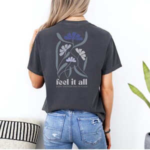 Puede incluir: Camiseta gris oscuro con un dise&ntilde;o floral en tonos azules y grises en la espalda. El texto "feel it all" est&aacute; debajo del dise&ntilde;o floral, con la frase "every emotion has its place" debajo.
