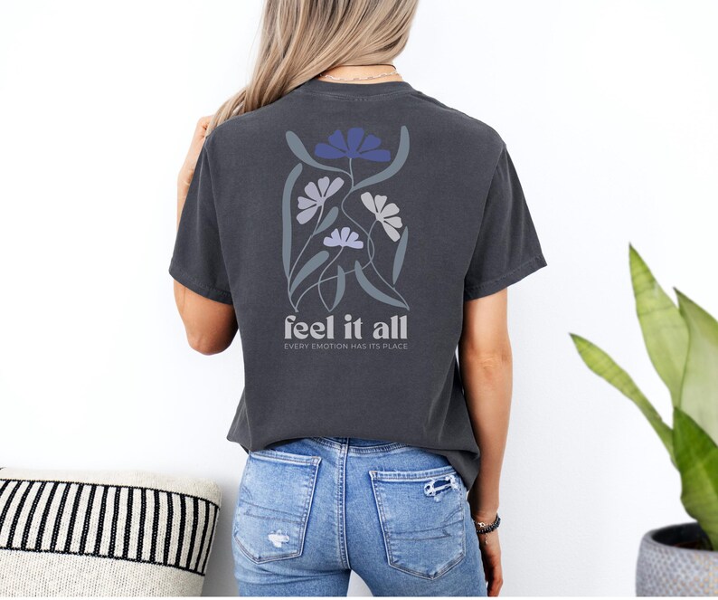 Puede incluir: Camiseta gris oscuro con un dise&ntilde;o floral en tonos azules y grises en la espalda. El texto "feel it all" est&aacute; debajo del dise&ntilde;o floral, con la frase "every emotion has its place" debajo.