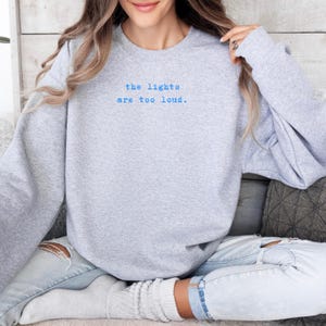 May include: A light gray sweatshirt with the text "the lights are too loud" in blue. The long-sleeved crew neck sweatshirt is made of a soft material. The sweatshirt is a casual style.