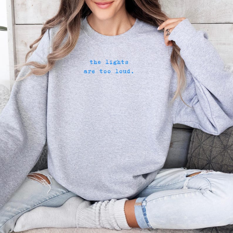 May include: A light gray sweatshirt with the text "the lights are too loud" in blue. The long-sleeved crew neck sweatshirt is made of a soft material. The sweatshirt is a casual style.