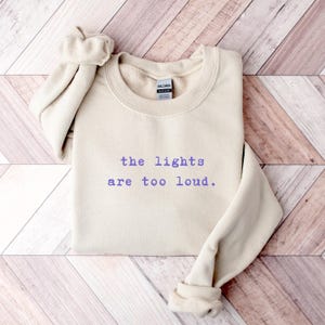 May include: A beige sweatshirt with the text "the lights are too loud" in purple. The long sleeves are folded up. The sweatshirt is lying on a wooden surface.