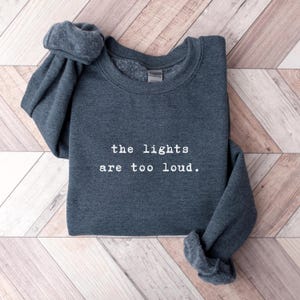 May include: A dark gray sweatshirt with the text "the lights are too loud" in white. The sweatshirt is folded with the sleeves rolled up. The background is a light brown wooden surface.
