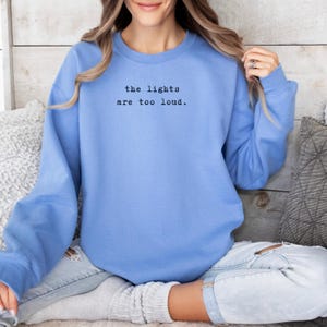 May include: A light blue sweatshirt with the text "the lights are too loud" in black. The long-sleeved top is made of a soft material and is suitable for casual wear.