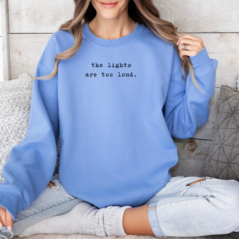 May include: A light blue sweatshirt with the text "the lights are too loud" in black. The long-sleeved top is made of a soft material and is suitable for casual wear.