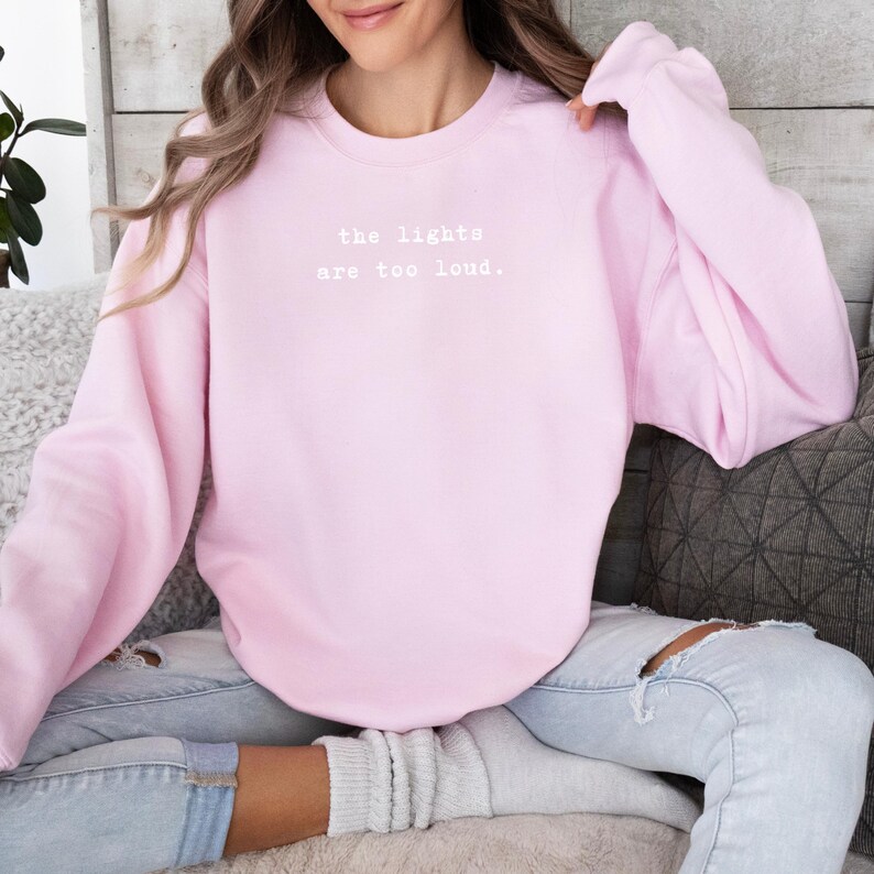 May include: A light pink sweatshirt with the text "the lights are too loud" in white. The long-sleeved crew neck sweatshirt is made of a soft material. The model is wearing light blue jeans.