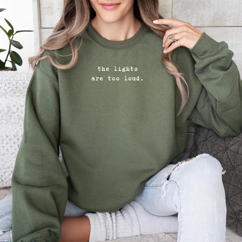 May include: An olive green sweatshirt with the text "the lights are too loud" in a white, simple font. The sweatshirt is a solid color and has a crew neck. The model is wearing light wash jeans.