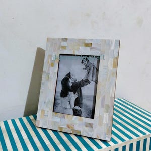 May include: A rectangular photo frame with a mosaic-style border of white and gold pieces. The frame holds a black and white photograph. The frame is resting on a blue and white striped surface.