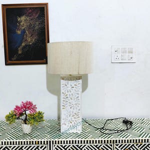 May include: A table lamp with a beige lampshade and a white, leaf-patterned base. The lamp is on a cabinet with a black and white geometric design. A framed horse painting hangs on the wall.