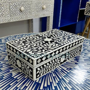 May include: A rectangular black and white decorative box with a floral design sits on a blue and white patterned table. The box features a star and leaf pattern on the lid. The table has a sunburst design. Other furniture is visible in the background.