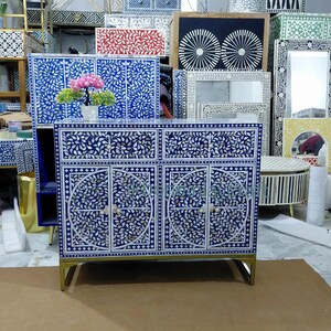 May include: A blue cabinet with a floral design made of white inlay. The cabinet has four doors and two drawers, with gold-coloured legs. A vase of pink flowers sits on top. Other furniture with similar designs is in the background.