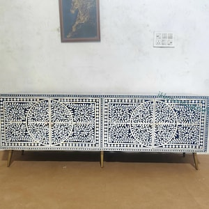 May include: A long, rectangular sideboard cabinet featuring a dark blue and white floral inlay pattern. The piece has four cabinet doors, each decorated with circular motifs and leaf designs, supported by four slender, tapered gold-toned metal legs.