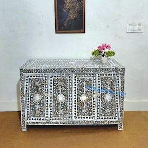 Puede incluir: Un aparador decorativo de cuatro puertas, con un diseño floral en blanco y negro. El mueble tiene forma rectangular y está adornado con intrincados diseños incrustados. Una obra de arte enmarcada y un pequeño arreglo floral se encuentran encima.