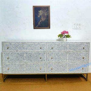 Op de afbeelding: Grijze dressoir met een bloemmotief en goudkleurige handgrepen. De dressoir heeft negen laden en staat op een goudkleurig metalen frame. Een ingelijste foto en een kleine plant staan erop.