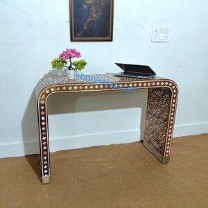 May include: A brown desk with a curved design, inlaid with white and brown decorative patterns. The desk has a rectangular top and a matching patterned front. A laptop sits on the desk, and a small potted plant is on the left side.