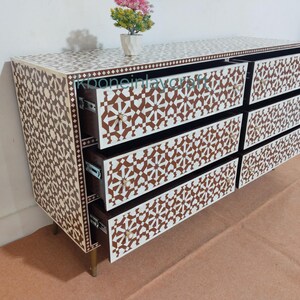 May include: A six-drawer chest of drawers with a geometric pattern in brown and white. The chest has a rectangular shape with gold-coloured legs and drawer pulls. A small vase with flowers sits on top.
