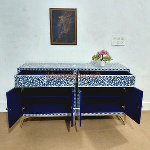 Puede incluir: Un aparador azul marino con dos puertas abiertas y dos cajones. El mueble está decorado con un estampado floral blanco y tiene patas doradas. Una imagen enmarcada y una pequeña planta se encuentran encima del aparador.