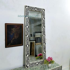 May include: A long, rectangular mirror with a black and white floral frame. The mirror is above a matching cabinet with a geometric pattern. A framed artwork and a small vase of pink flowers are also visible.