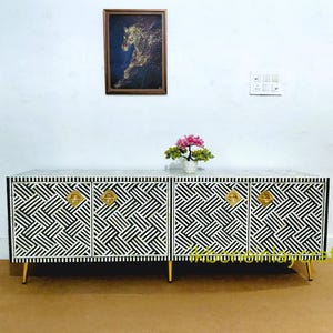 Puede incluir: Un mueble con estampado en blanco y negro con herrajes y patas doradas. El mueble tiene cuatro puertas con un diseño geométrico. Una imagen enmarcada de un caballo y un pequeño arreglo floral encima.