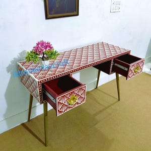 May include: A patterned desk with a red and white geometric design. It features two drawers with gold handles and slender gold legs. The desk has a small plant on top. The desk's dimensions are approximately 120 cm in length.