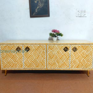 May include: A yellow and white patterned cabinet with four doors and brass hardware. The cabinet has a geometric design and sits on thin, gold-coloured legs. A small potted plant with pink flowers sits on top.