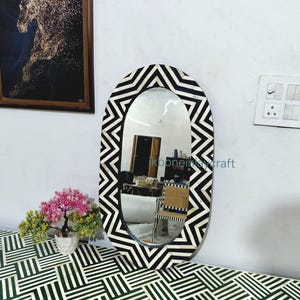 Puede incluir: Espejo ovalado con marco de patrón de chevrón blanco y negro. El espejo se exhibe sobre una mesa con un diseño geométrico verde y blanco. Una obra de arte enmarcada de un caballo cuelga en la pared.