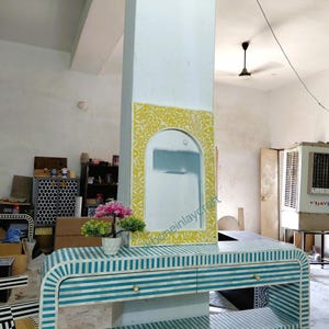 Puede incluir: Una consola moderna con un diseño de rayas azules y blancas. La mesa tiene dos cajones y un frente curvo. Un espejo con un marco floral amarillo está montado en un pilar blanco. Una pequeña planta en maceta se encuentra sobre la mesa. La habitación tiene una estética de taller.