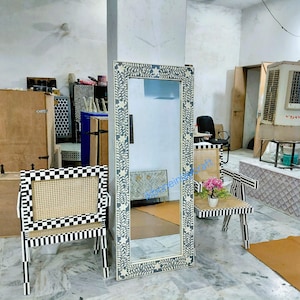 May include: A tall, rectangular mirror with a grey and white floral border. Two chairs with black and white chequered and striped frames and woven cane seats are in the foreground. A small table with a striped frame and woven cane top is also visible.
